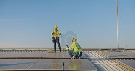 ingénieur Techniciens solaires en vestes haute visibilité et casques de sécurité communique à l'aide walkie-talkie tandis qu'un autre siège avec une tablette sur un tableau de panneaux solaires sur une ferme solaire sur le toit sous un clair