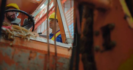 Trabajadores de la construcción en sombreros duros amarillos y chalecos de seguridad operan una maquinaria de elevación de construcción dentro de un entorno industrial, se centran en la seguridad ocupacional y el trabajo en equipo