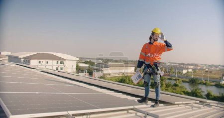 A solar panel technician engineer in safety harness and hard hat inspects a rooftop solar installation with a clipboard in hand against a clear sky and industrial landscape