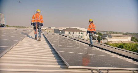 Solar panel engineers in orange high-visibility jackets and safety helmets walk on a metal roof, inspecting the newly installed solar system