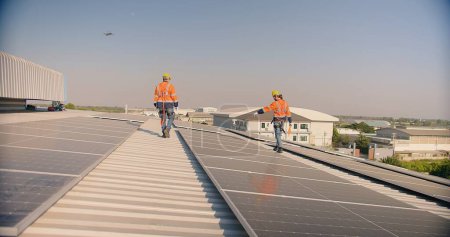Photo for Solar panel engineers in orange high-visibility jackets and safety helmets walk on a metal roof, inspecting the newly installed solar system - Royalty Free Image