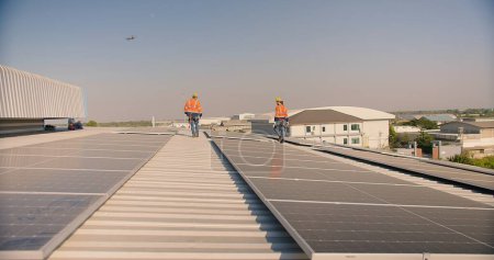 Solar panel engineers in orange high-visibility jackets and safety helmets walk on a metal roof, inspecting the newly installed solar system