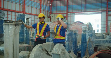 Trabajadores industriales con tableta y auriculares en almacén, evalúan maquinaria envuelta en plástico protector en un almacén industrial durante un control de calidad.