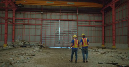 Two construction workers in yellow hard hats and reflective vests discussing stand amidst a large industrial site, examine a building site with structural steelwork in the background