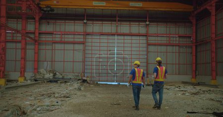 Two construction workers in yellow hard hats and reflective vests discussing stand amidst a large industrial site, examine a building site with structural steelwork in the background