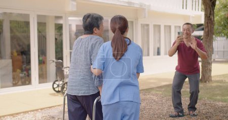 Enfermera asiática que apoya el cuidado de la paciente adulta al aire libre durante la recuperación de las lesiones de las piernas y el marido anciano filma video de su esposa, enfermera que empodera a los pacientes ancianos en su recuperación