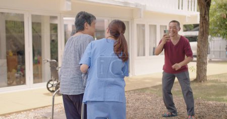 Asiatische Krankenschwester unterstützt die Pflege erwachsener Patientinnen im Freien während der Genesung von Beinverletzungen und älterer Ehemann filmt Video seiner Frau, Krankenschwester unterstützt ältere Patienten bei ihrer Genesung