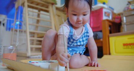 Un bambin absorbé dans une robe à carreaux bleus découvre la créativité avec un pinceau à la main et une palette de peinture colorée sur le sol, tout-petit aime la peinture, l'activité mêlant jeu et créativité
