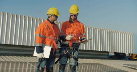 Two solar engineers in reflective gear discuss work plans project details on a rooftop with solar panels