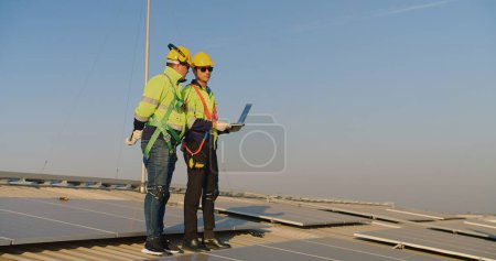 Two engineers in safety gear are discussing over a laptop on a large solar panel field during a sunny day.