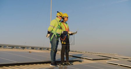 Two engineers in safety gear are discussing over a laptop on a large solar panel field during a sunny day.