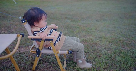 Ein neugieriges Kleinkind, das auf einem Klappstuhl hockt, wendet sich zum Wegschauen und ruft ein Gefühl des Staunens hervor, das sich gegen die weichen Farbtöne einer Rasenfläche richtet.