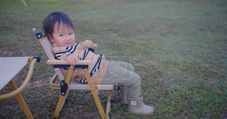 A curious toddler perched on a foldable chair turns to look away, evoking a sense of wonder, set against the soft hues of a grassy field