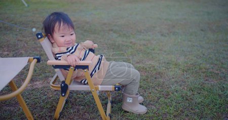 A curious toddler perched on a foldable chair turns to look away, evoking a sense of wonder, set against the soft hues of a grassy field