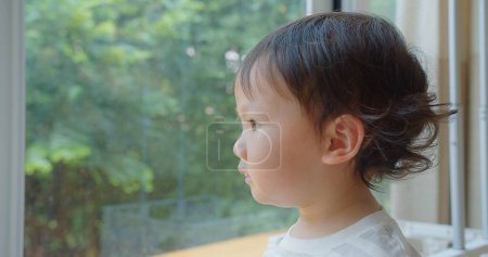 Ein nachdenkliches kleines Kind blickt aus einem Fenster in einen üppigen Garten, verloren in Gedanken, mit sanftem natürlichem Licht, das einen friedlichen Ausdruck hervorhebt
