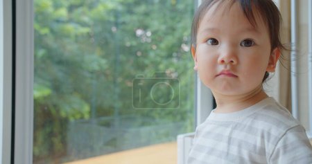 Un jeune enfant cher regarde par une fenêtre dans un jardin luxuriant, perdu dans la pensée, avec une lumière naturelle douce accentuant une expression paisible