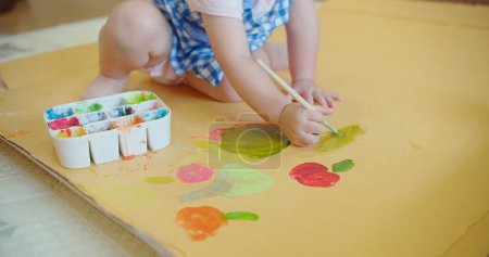 An engrossed toddler in a blue checkered dress discovers creativity with a paintbrush in hand and colorful paint palette on the floor, toddler enjoys painting, activity blending play with creativity