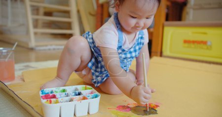 Un bambin absorbé dans une robe à carreaux bleus découvre la créativité avec un pinceau à la main et une palette de peinture colorée sur le sol, tout-petit aime la peinture, l'activité mêlant jeu et créativité