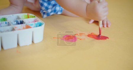 An engrossed toddler in a blue checkered dress discovers creativity with a paintbrush in hand and colorful paint palette on the floor, toddler enjoys painting, activity blending play with creativity