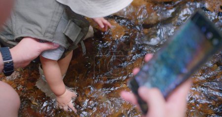 A small child explores a clear mountain stream, guided by parent helping hand, with the moment being captured on a smartphone