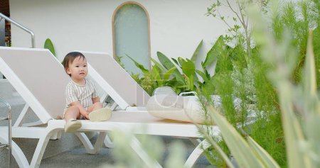 Ein Kleinkind sitzt zufrieden auf einer weißen Sonnenliege inmitten üppigen Grüns und verkörpert eine heitere tropische Atmosphäre