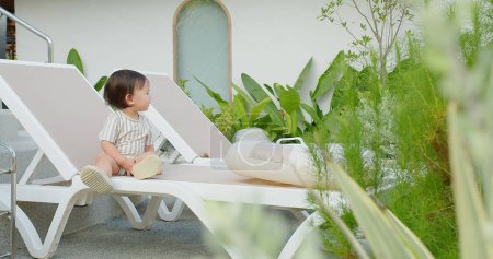 A toddler sits contentedly on a white sun lounger surrounded by lush greenery, embodying a serene tropical atmosphere