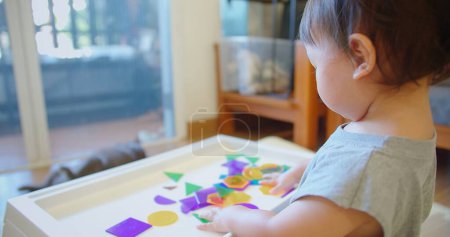 Über-die-Schulter-Ansicht eines Kleinkindes, das mit transluzenten farbigen Formen auf einem hellen Tisch spielt und eine helle, einnehmende Aktivität zeigt, die die sensorische und kognitive Entwicklung anregt