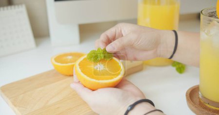 A person is cutting an orange and placing a leaf on it. There is a glass of orange juice on the table