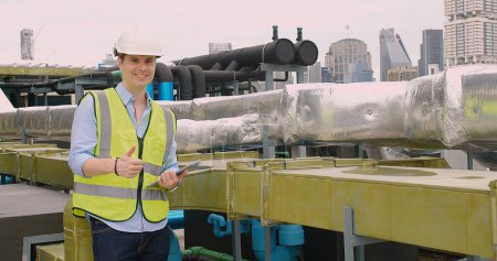 Smiling engineer giving thumbs up on a rooftop with insulated piping and cityscape in the background.
