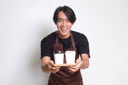 Téléchargez les photos : Portrait d'homme barista asiatique attrayant en tablier brun tenant deux tasses à café en papier à emporter. Image isolée sur fond blanc - en image libre de droit