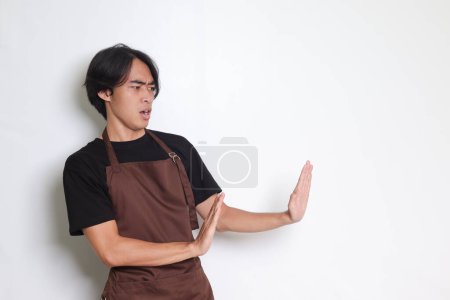 Téléchargez les photos : Portrait d'homme barista asiatique attrayant en tablier brun formant un geste de main pour éviter quelque chose. Image isolée sur fond blanc - en image libre de droit