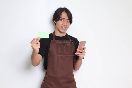 Téléchargez les photos : Portrait d'homme barista asiatique attrayant en tablier brun montrant modèle de carte de crédit et pointant sur elle. Image isolée sur fond blanc - en image libre de droit