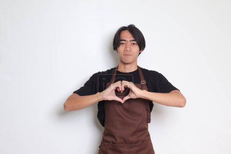 Téléchargez les photos : Portrait de barista asiatique attrayant homme en tablier brun parle de ses propres sentiments, fait geste de cœur sur la poitrine, exprime la sympathie et l'amour, sourit positivement. Image isolée sur fond blanc - en image libre de droit