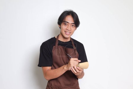 Téléchargez les photos : Portrait d'homme barista asiatique attrayant en tablier brun prenant l'ordre, l'écriture sur la liste des livres de menu. Image isolée sur fond blanc - en image libre de droit