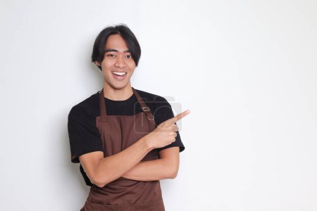 Téléchargez les photos : Portrait d'homme barista asiatique attrayant en tablier brun montrant le produit, pointant quelque chose avec les mains. Concept publicitaire. Image isolée sur fond blanc - en image libre de droit