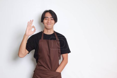 Téléchargez les photos : Portrait d'homme barista asiatique attrayant en tablier brun montrant ok geste de la main et souriant en regardant la caméra. Concept publicitaire. Image isolée sur fond blanc - en image libre de droit