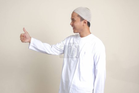 Téléchargez les photos : Portrait de l'homme musulman asiatique attrayant en chemise blanche montrant pouce vers le haut, bon geste de la main du travail. Image isolée sur fond gris - en image libre de droit
