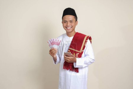 Téléchargez les photos : Portrait de l'homme musulman asiatique attrayant en chemise blanche montrant cent mille rupiah tout en utilisant le téléphone mobile. Concept financier et d'épargne. Image isolée sur fond gris - en image libre de droit