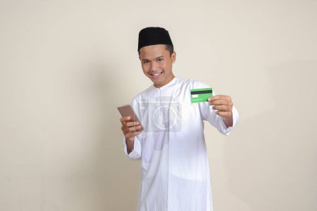 Téléchargez les photos : Portrait d'homme musulman asiatique attrayant en chemise blanche avec un bonnet de crâne tenant un téléphone portable et présentant une carte de crédit. Image isolée sur fond gris - en image libre de droit