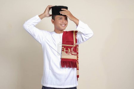 Téléchargez les photos : Portrait d'homme musulman asiatique séduisant en chemise blanche avec calotte crânienne essayant d'ajuster sa calotte crânienne chantée ou noire. Image isolée sur fond gris - en image libre de droit