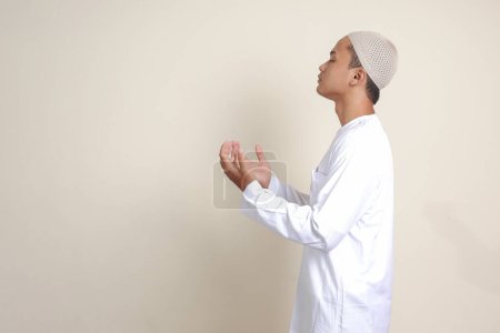 Téléchargez les photos : Portrait d'homme musulman asiatique attrayant en chemise blanche avec crâne priant sincèrement avec les mains levées. Image isolée sur fond gris - en image libre de droit