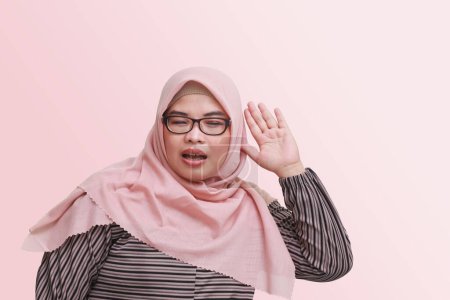 Téléchargez les photos : Portrait de femme asiatique joyeuse avec hijab, essayant d'entendre des commérages ou du son, tenant la main près de l'oreille et écoutant attentivement. Concept publicitaire. Image isolée sur fond rose - en image libre de droit