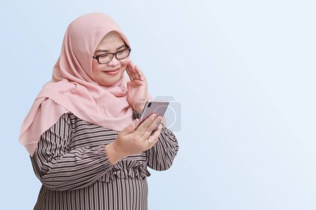 Téléchargez les photos : Portrait de femme asiatique joyeuse avec hijab, tenant le téléphone portable et prenant une photo d'elle-même ou selfie tandis que sa main touche la joue. Concept publicitaire. Image isolée sur fond bleu - en image libre de droit
