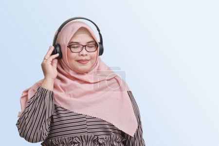 Téléchargez les photos : Portrait de femme asiatique joyeuse avec hijab, profiter et écouter de la musique à l'aide d'un casque. Image isolée sur fond bleu - en image libre de droit