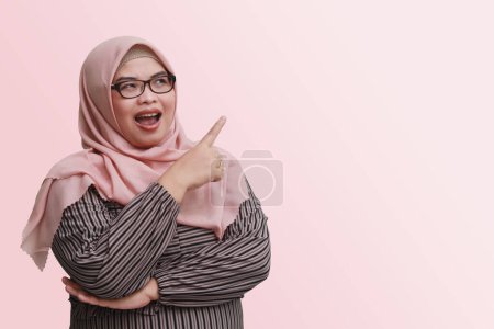Téléchargez les photos : Portrait de femme asiatique joyeuse avec hijab, montrant le produit, pointant quelque chose et souriant. Concept publicitaire. Image isolée sur fond rose - en image libre de droit