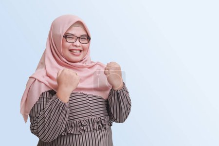 Téléchargez les photos : Portrait de femme asiatique joyeuse avec hijab, levant le poing, célébrant le succès. Concept publicitaire. Image isolée sur fond bleu - en image libre de droit