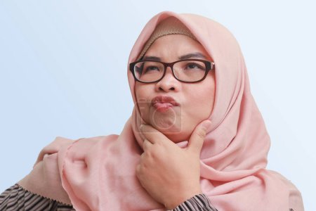 Téléchargez les photos : Portrait de femme asiatique joyeuse avec hijab, debout sur fond bleu, penser à la question avec la main sur le menton - en image libre de droit