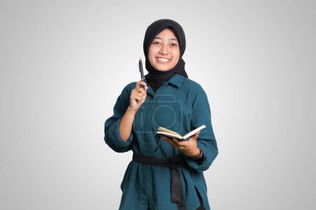 Téléchargez les photos : Portrait de femme musulmane asiatique excitée avec hijab écrit sur le carnet de notes et pointant vers le haut avec stylo. Concept publicitaire. Image isolée sur fond blanc - en image libre de droit