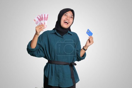 Photo for Portrait of cheerful Asian muslim woman with hijab, showing one hundred thousand rupiah while holding a credit card. Financial and savings concept. Isolated image on white background - Royalty Free Image