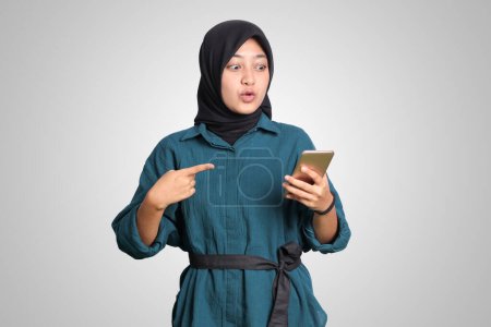 Téléchargez les photos : Portrait de femme musulmane asiatique excitée avec hijab tenant et touchant téléphone portable tout en pointant du doigt. Concept publicitaire. Image isolée sur fond blanc - en image libre de droit